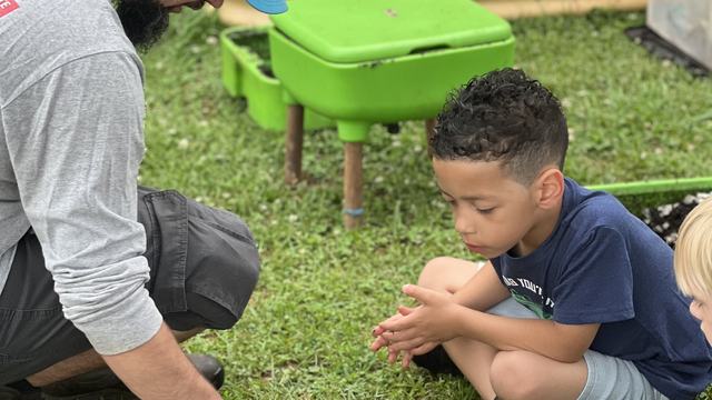 Man in blue hat squats down to show a child in a blue shirt the compost in a clear plastic plate. The child sits cross legged looking down at the worms, dirt, and egg shells. In the background are green worm composting bins and a raised bed for gardening.