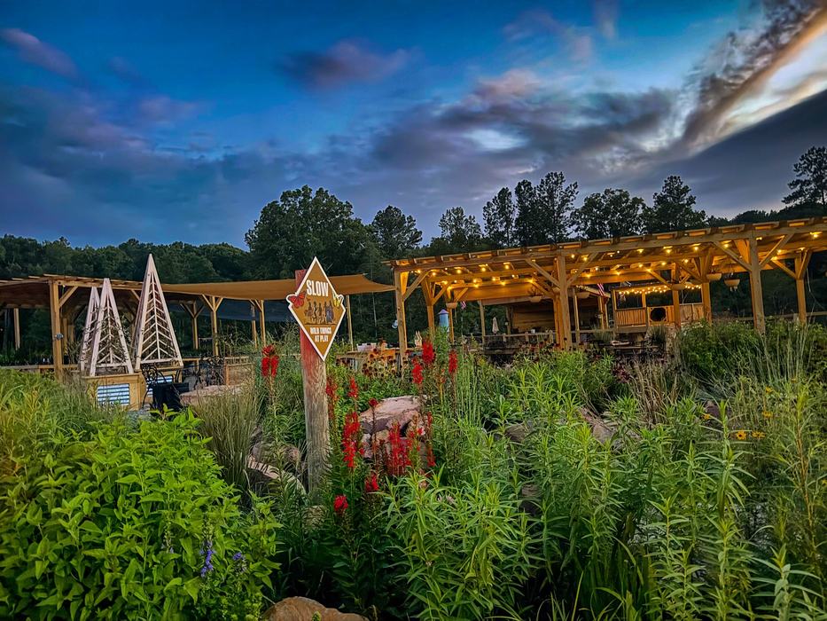 Garden with tall plants, "SLOW Wild Things At Play" sign and lit wooden pavilion at dusk