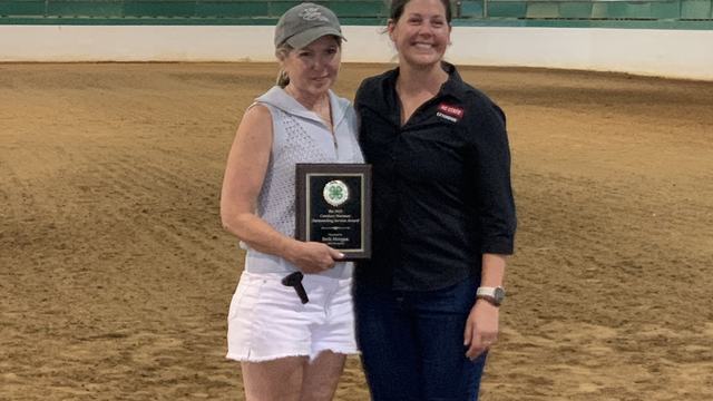 Two women standing in an indoor riding arena holding a 4‑H award plaque