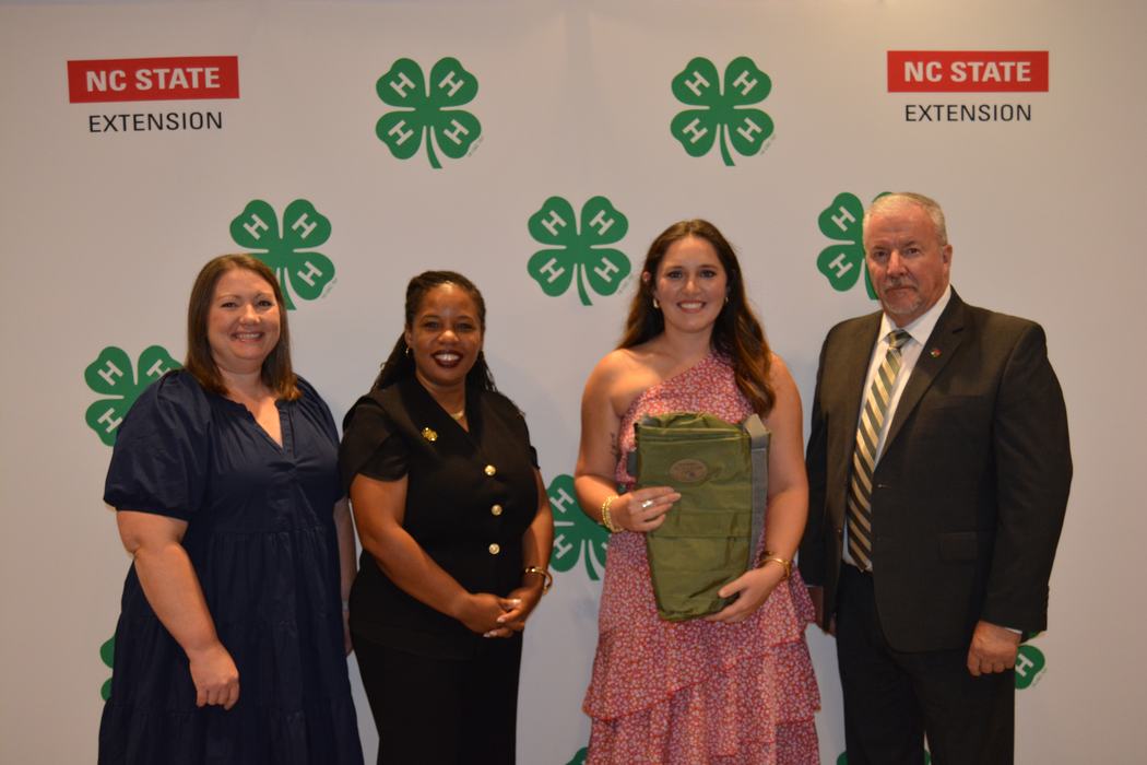 A woman holds her award while standing with others in front of a 4-H Banner.