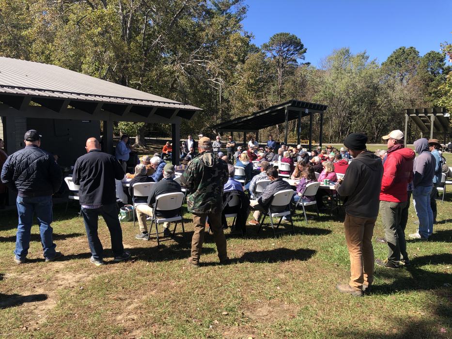 A large group of people near a picnic shelter all facing a person speaking.