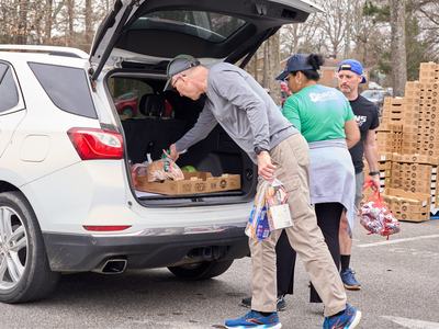 Volunteers loading groceries into open SUV trunk at distribution; shirt reads "PORCH"