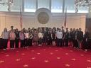 Large group of students and adults posed in a state legislative chamber with seal and flags