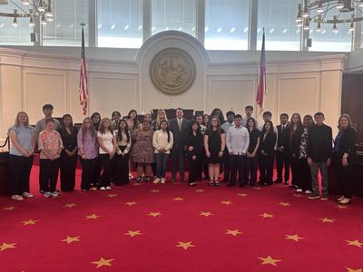 Large group of students and adults posed in a state legislative chamber with seal and flags