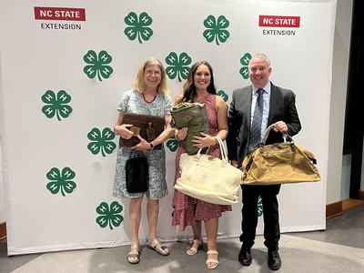 Three people holding bags in front of 4‑H clover backdrop with "NC STATE EXTENSION" text.