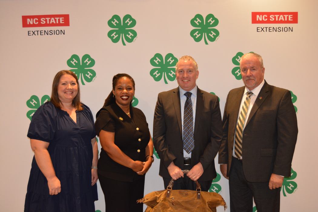 A county extension director holds an award while standing with others in front of a 4-H Banner.
