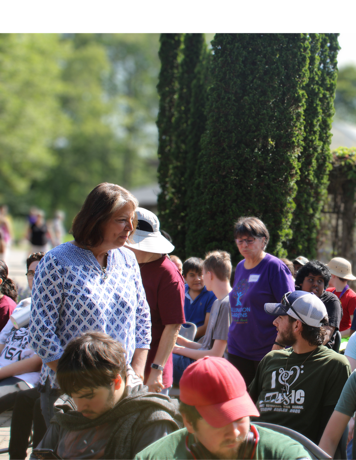 A middle-aged woman in a patterned blue and white shirt stands in a sunny outdoor crowd of diverse people, some seated, with tall cypress trees in the background.