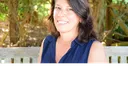 A smiling woman with dark hair and gray streaks sits on a wooden bench outdoors, wearing a navy blue sleeveless collared shirt. The background is filled with green foliage and trees.