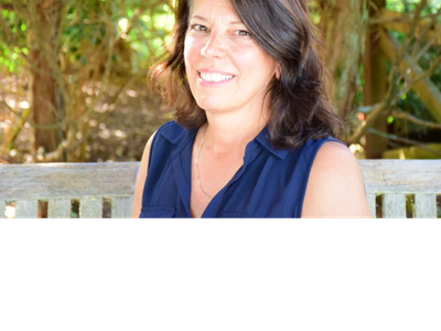 A smiling woman with dark hair and gray streaks sits on a wooden bench outdoors, wearing a navy blue sleeveless collared shirt. The background is filled with green foliage and trees.