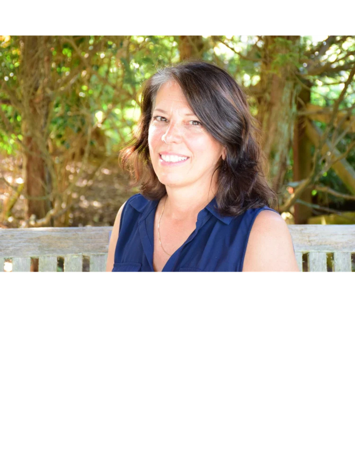 A smiling woman with dark hair and gray streaks sits on a wooden bench outdoors, wearing a navy blue sleeveless collared shirt. The background is filled with green foliage and trees.