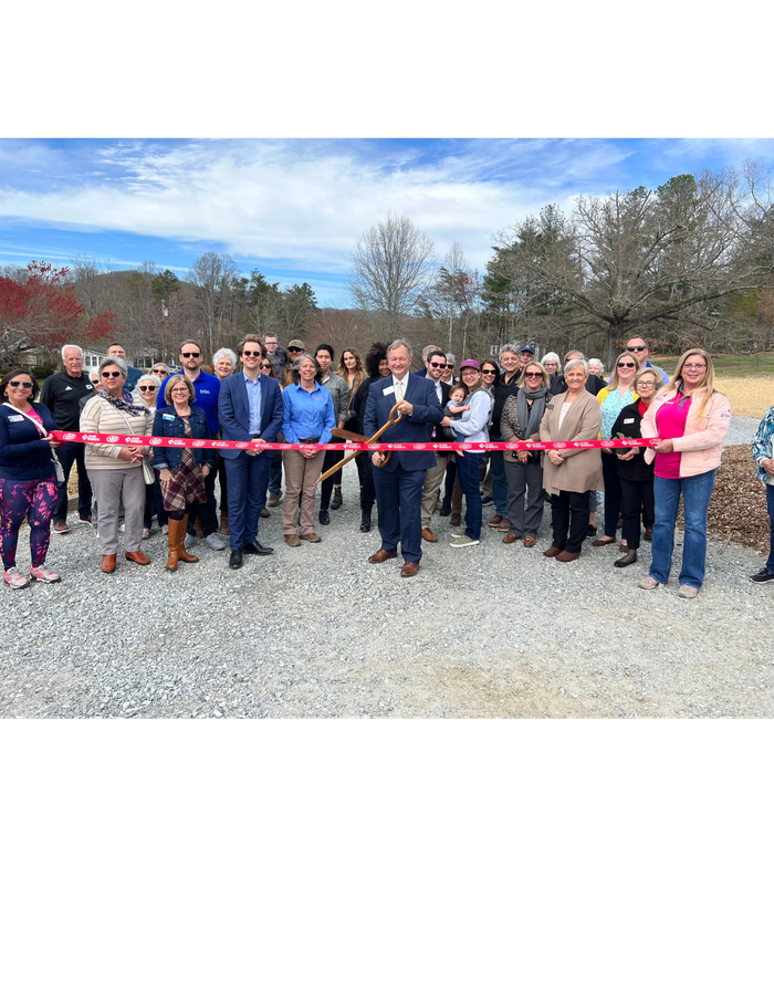 A diverse group of around 40 people stands outside on a gravel path, smiling for a photo behind a red ribbon. A man in a dark suit in the center holds a large pair of scissors, ready for a ribbon-cutting ceremony. Trees and hills are in the background under a blue sky.