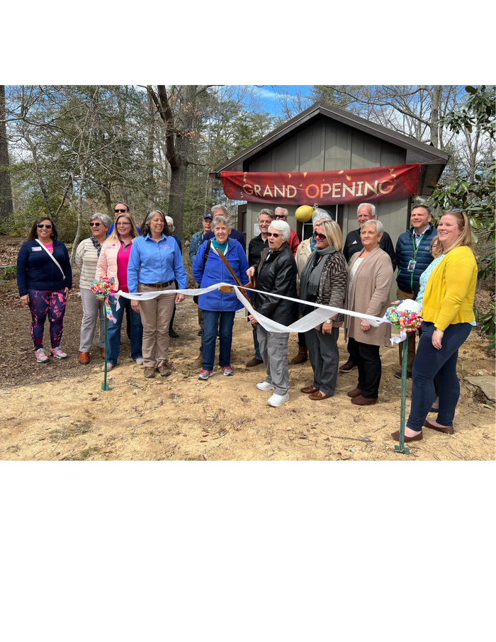 A group of about 15 adults stand outdoors in front of a small cabin with a banner reading "GRAND OPENING" for a ribbon-cutting ceremony. One woman in the center holds the scissors, preparing to cut the ribbon. The ground is dirt and the background is a wooded area.