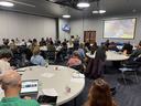 Conference room with attendees seated at round tables listening to a presenter at front