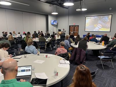 Conference room with attendees seated at round tables listening to a presenter at front