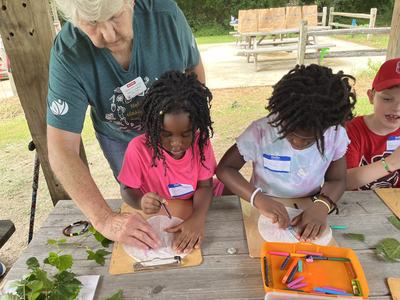 A woman helps children draw leaves at an outdoor classroom.