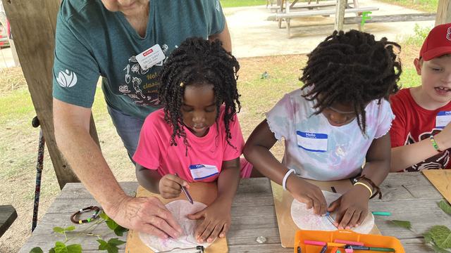 A woman helps children draw leaves at an outdoor classroom.