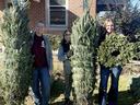 Three people standing in a yard holding wrapped Christmas trees and a wreath