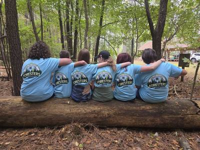 Six people sitting on a log with arms around each other wearing blue shirts labeled Foresters 2025