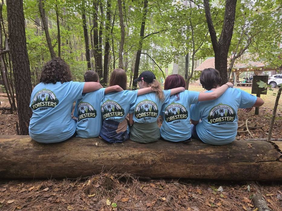 Six 4-H youth sit on a log in the forest with their backs to the camera, showing the text on their shirts that reads Onslow County Foresters 2025