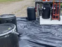 Black plastic barrels on a black tarp beside a truck and an orange-vested hand truck