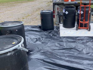 Black plastic barrels on a black tarp beside a truck and an orange-vested hand truck