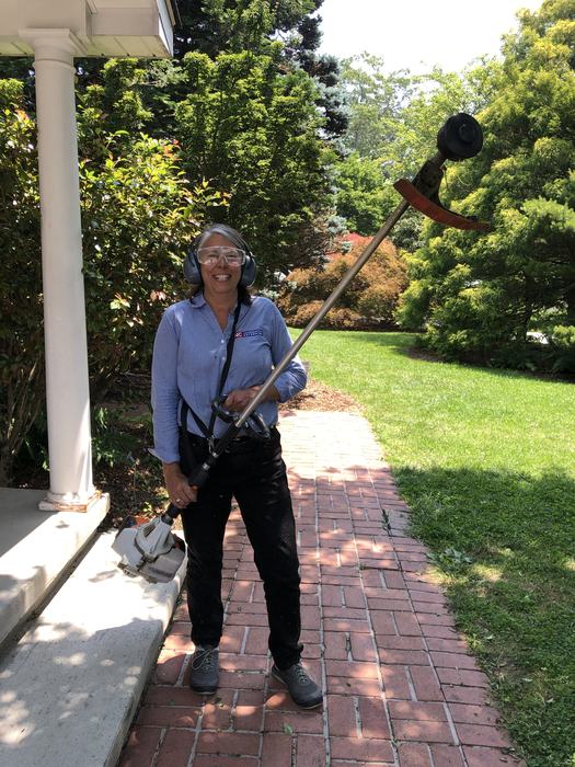 A smiling woman wearing a blue and white striped shirt, safety glasses, and ear protection stands on a brick path holding a weed trimmer.