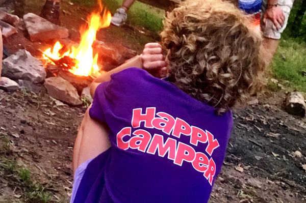 A child sits beside a campfire in a shirt that reads "Happy Camper".