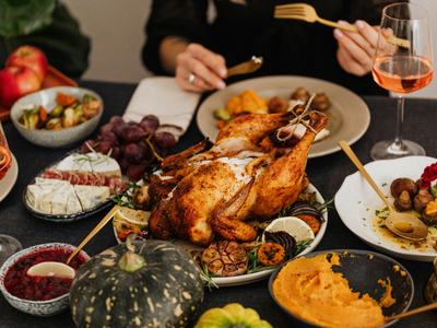 Photo of a table of fall decor and various foods. A roasted turkey sits at the center of the table. In the background you see the hands of someone holding eating utensils over a plate.