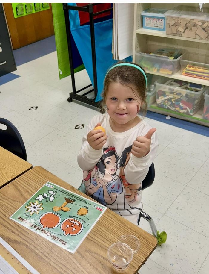 a little girl giving a thumbs up sign with a pumpkin life cycle mat on her desk