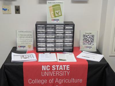 table with red banner, small parts cabinet containing seeds, paper materials and resources on table, white wall behind table
