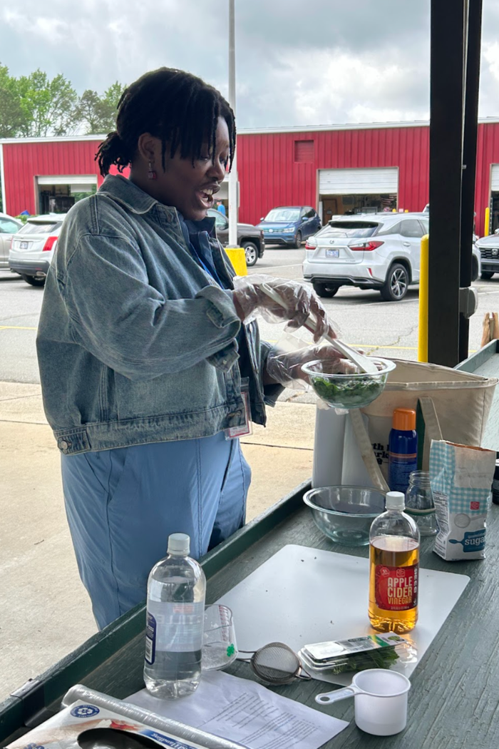 Person in denim jacket stirring salad in plastic bowl at outdoor table; bottle labeled "Apple Cider Vinegar"