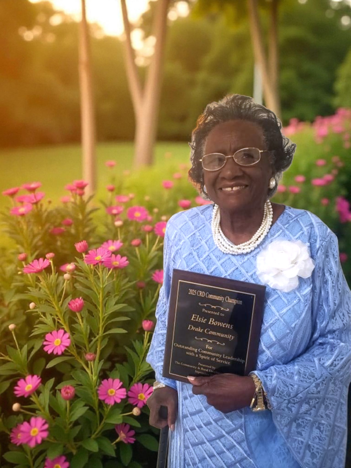 Elderly woman in blue dress holding a "2025 CRD Community Champion" plaque beside pink flowers
