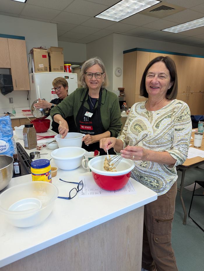 Three women in a kitchen stirring mixing bowls, one wearing an NC STATE apron
