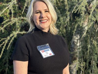 A smiling blonde woman wearing a black top and a name badge stands in front of a leafy tree.