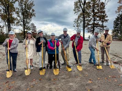 Nine people in hard hats holding gold shovels at a lakeside groundbreaking