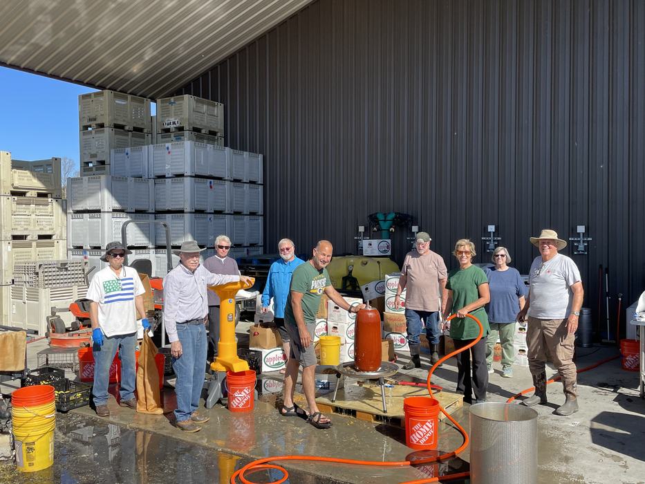 Group of people making cider or juice outside a storage building with crates.