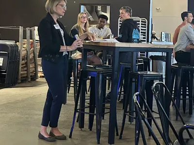 Woman speaking to seated group at high table in bar; TV above shows "Bojangles" baseball ad