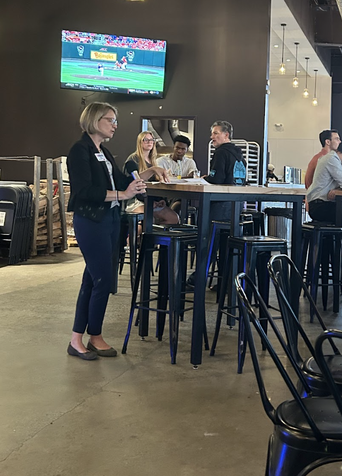 Woman speaking to seated group at high table in bar; TV above shows "Bojangles" baseball ad
