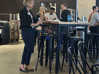 Woman speaking to seated group at high table in bar; TV above shows "Bojangles" baseball ad