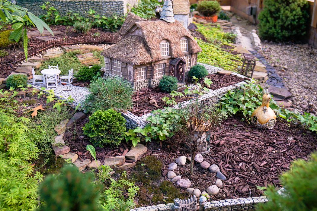 A scene of miniature plants and wall around a miniature house. Tiny garden furniture is visible, as is a path and arbor.
