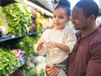 Father holding daughter near produce aisle; text "Produce in Season"