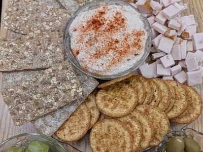 Platter with dip sprinkled with paprika, crispbread, round crackers, cubed ham, grapes, olives