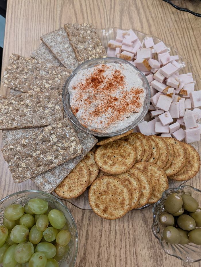 Platter with dip sprinkled with paprika, crispbread, round crackers, cubed ham, grapes, olives