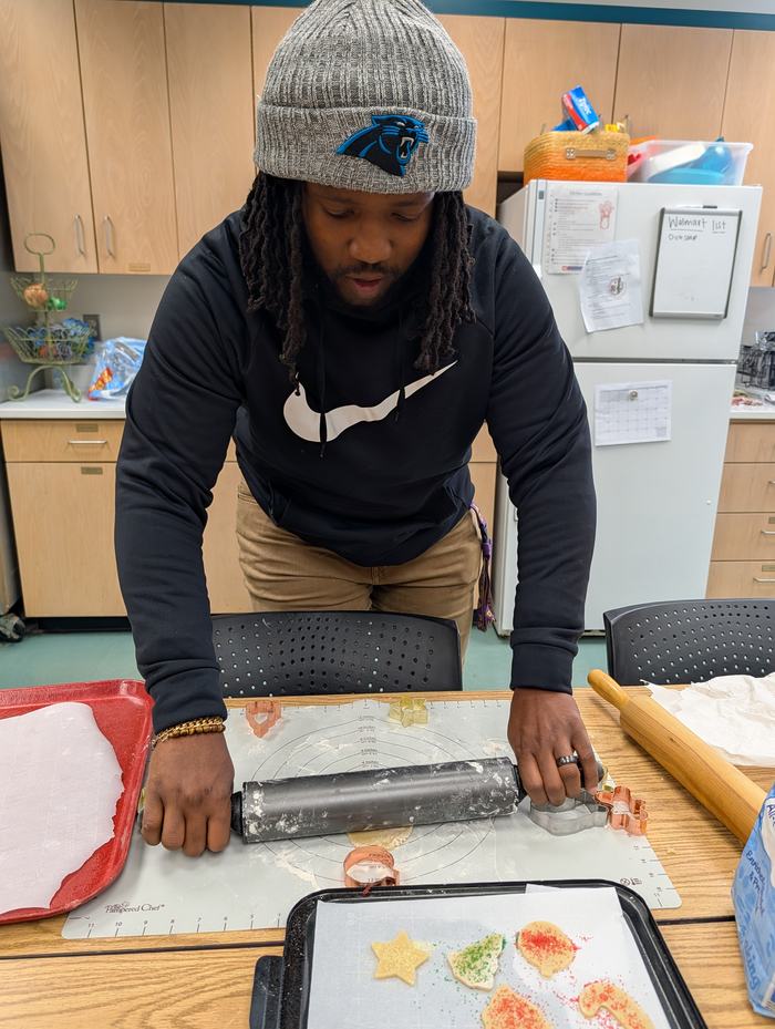 Man wearing beanie rolling cookie dough with rolling pin and cookie cutters on mat