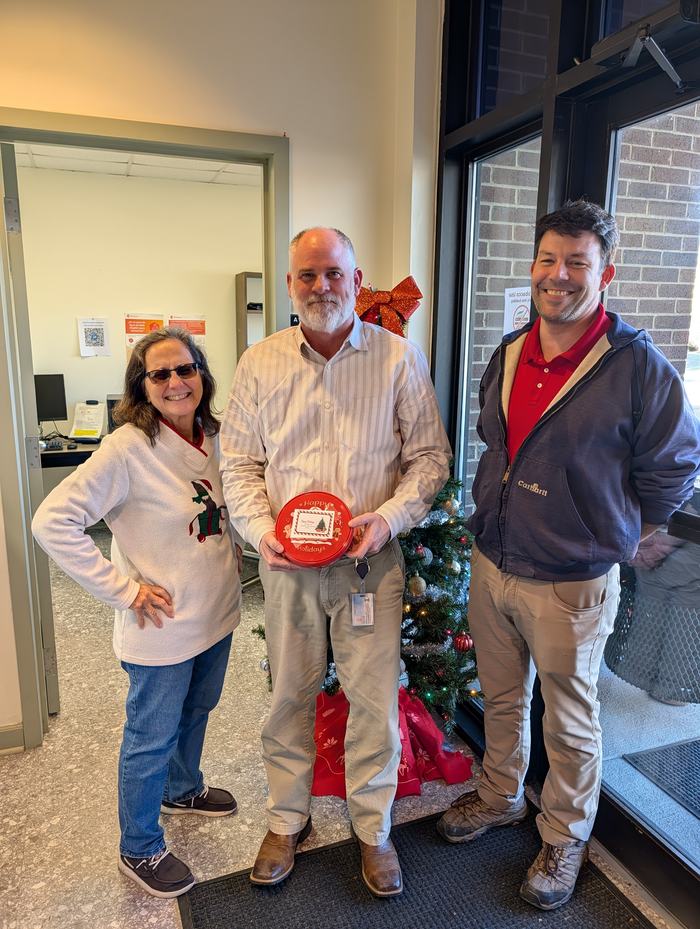 Three people standing by a small Christmas tree; center person holds red tin reading "Happy Holidays"