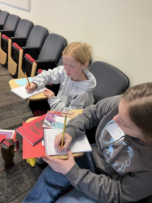 Two students seated in auditorium chairs writing in notebooks on fold-down desks