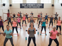 An indoor fitness class that consists of a diverse group of men and women of various ages and fitness levels, all performing a strength training exercise with light weights (dumbbells) and resistance bands. The participants are arranged in rows, facing th