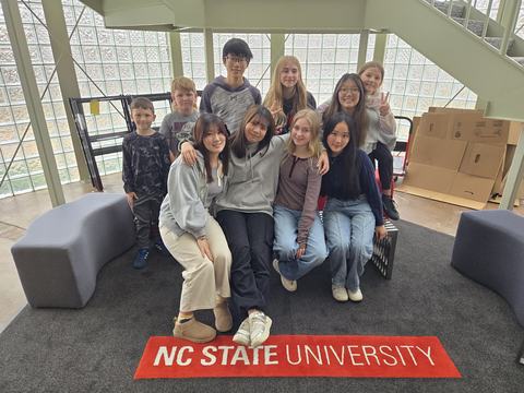 A large group of kids and teenagers are grouped together, posing for the photo. The carpet near their feet is printed with "NC State University." They were the AYP students and youth of the host families in 2024-2025.