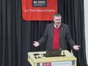 A man in a suit and red sweater presents at a podium with "N.C. Plant Sciences Initiative" signage. He gestures openly, conveying an engaging speech.