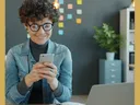 Person with curly hair and glasses using smartphone at desk with laptop, notebook, sticky notes